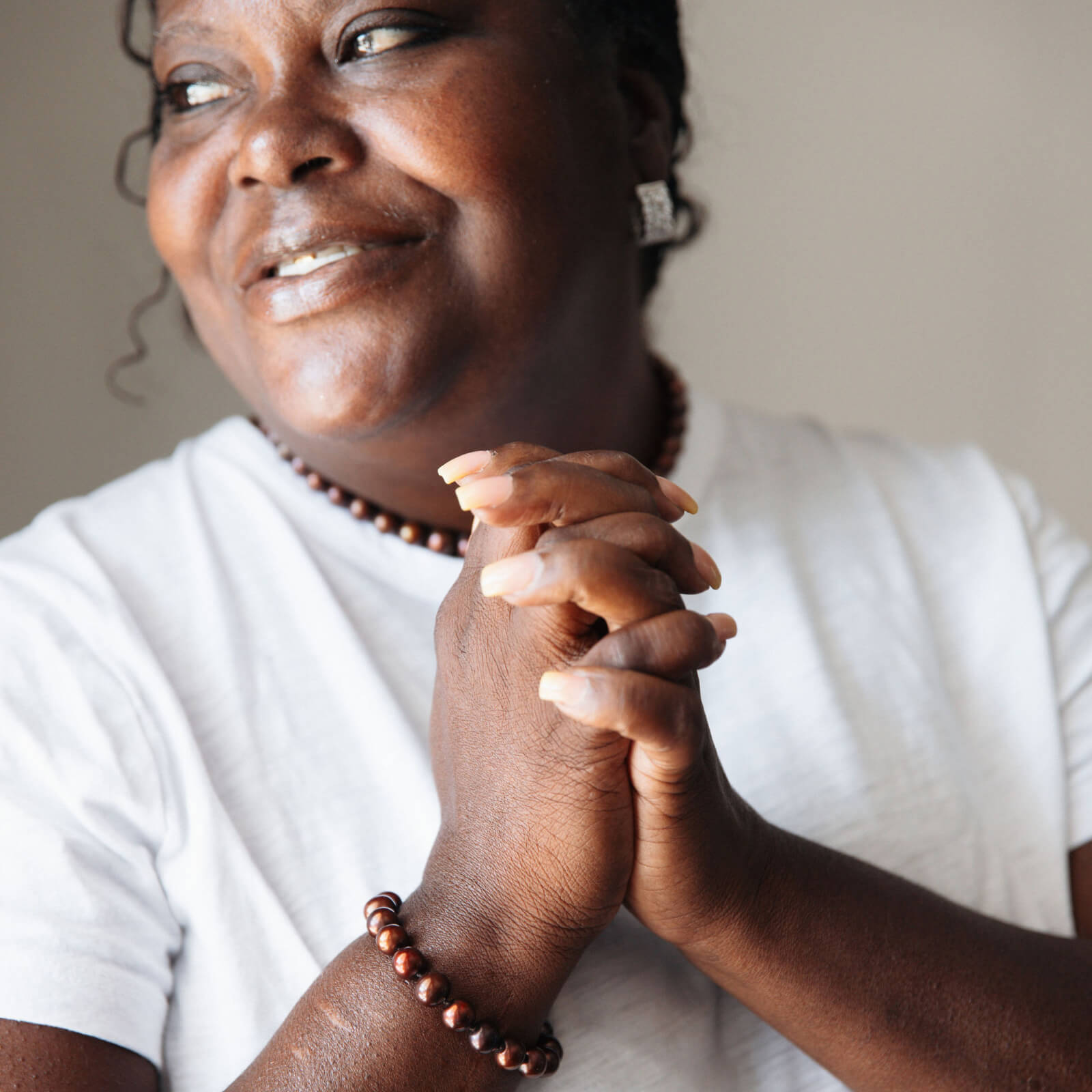 Woman with clasped hands wearing a white shirt and beaded bracelet against a neutral background