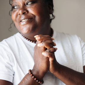 Woman with clasped hands wearing a white shirt and beaded bracelet against a neutral background