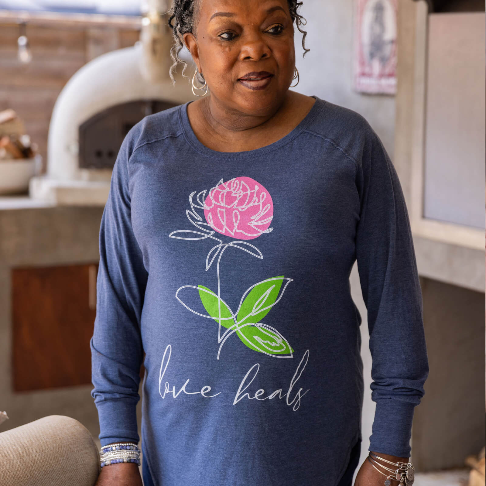 Woman wearing a blue long-sleeve shirt with a pink flower and 'love heals' text in a kitchen.