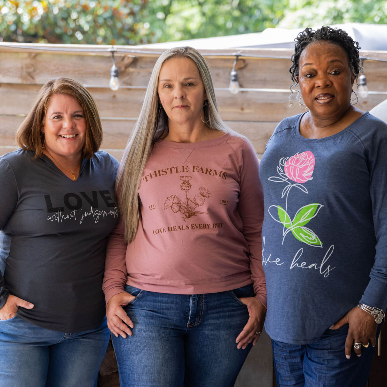Three women standing together wearing Thistle Farms branded shirts with visible text and graphics.