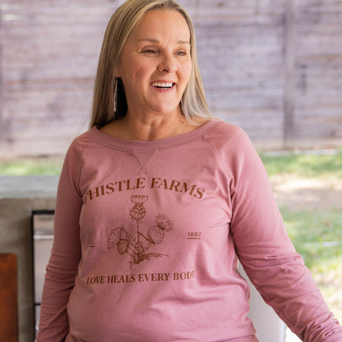Woman wearing a pink long-sleeve shirt with 'Thistle Farms' branding and text.