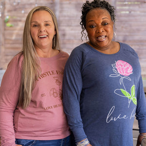 Two women wearing pink and blue Thistle Farms branded shirts with floral designs and text.