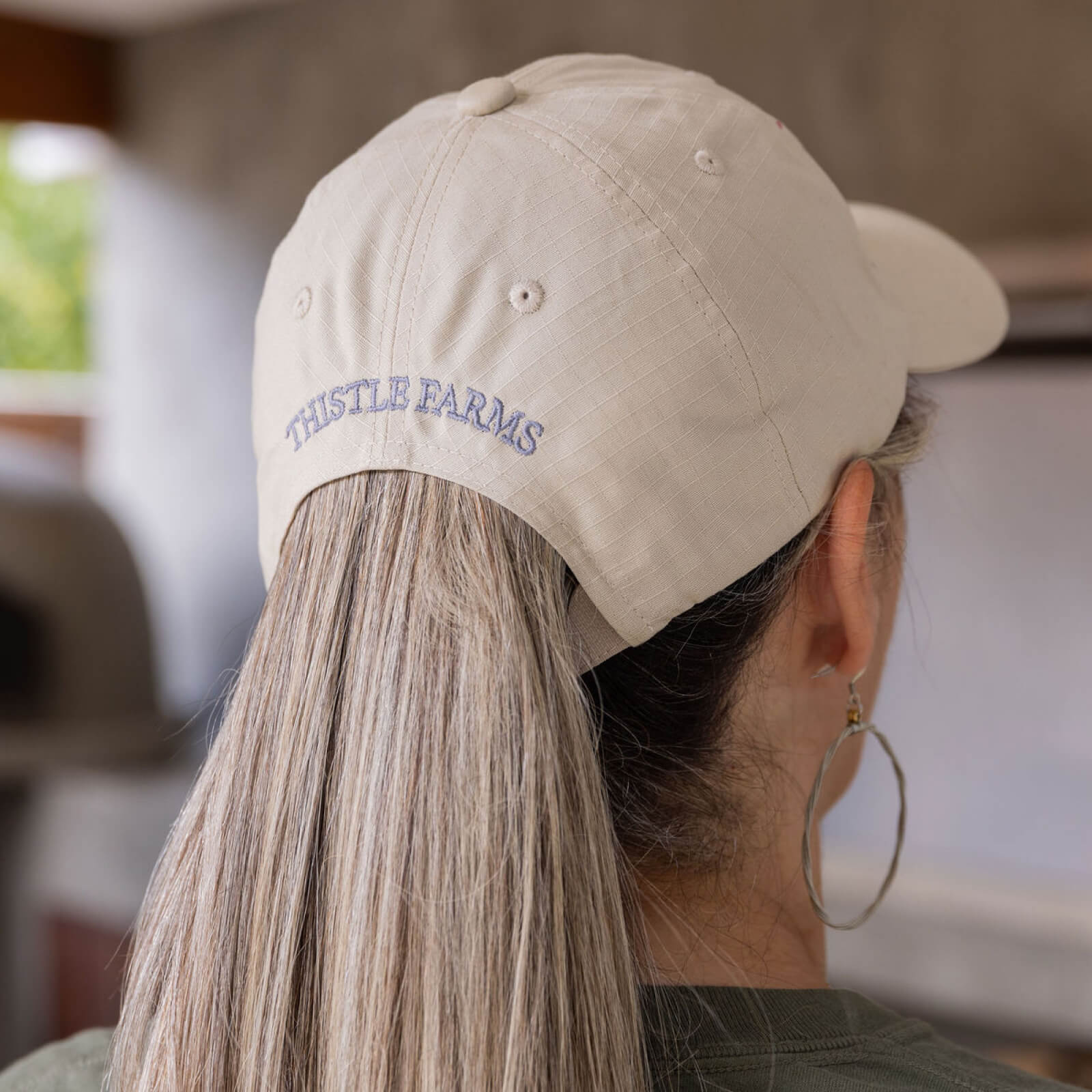 Person wearing a beige cap with 'Thistle Farms' logo, blurred background
