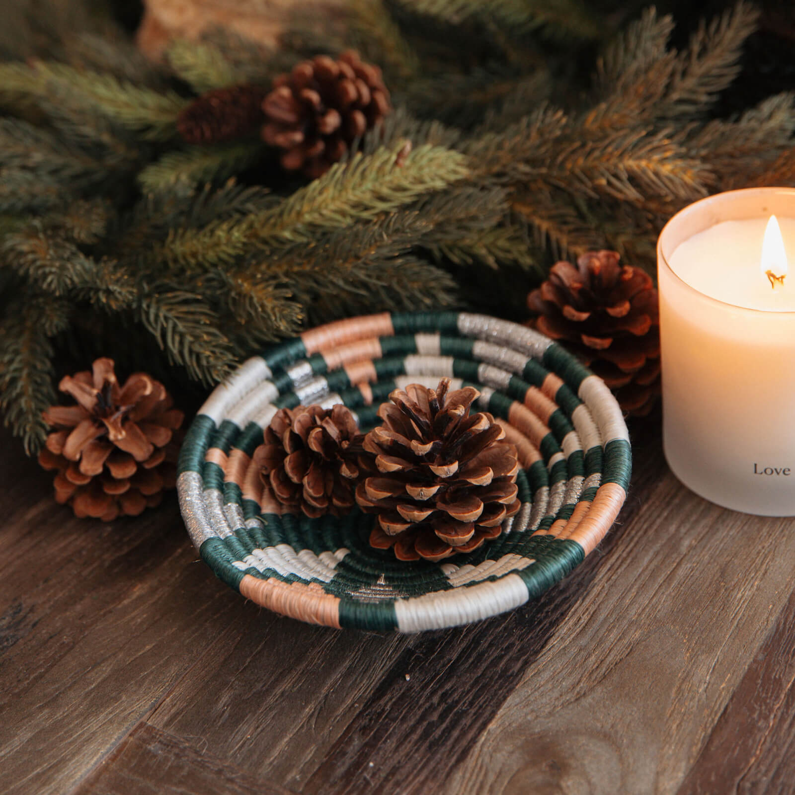 Woven basket with pine cones on a wooden surface next to a lit candle.
