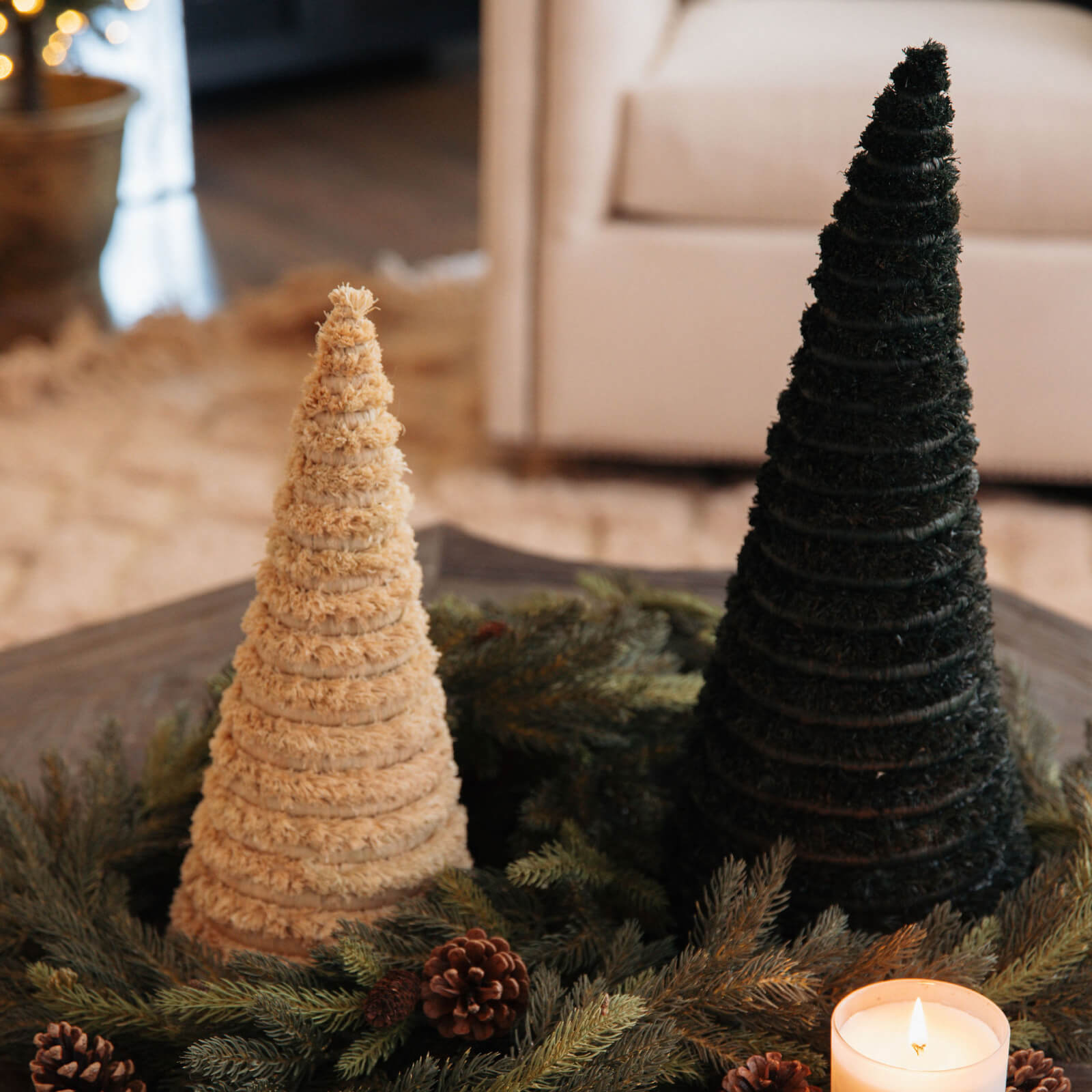 Two decorative Christmas trees, one beige and one dark green, on a table with a candle and pinecones.