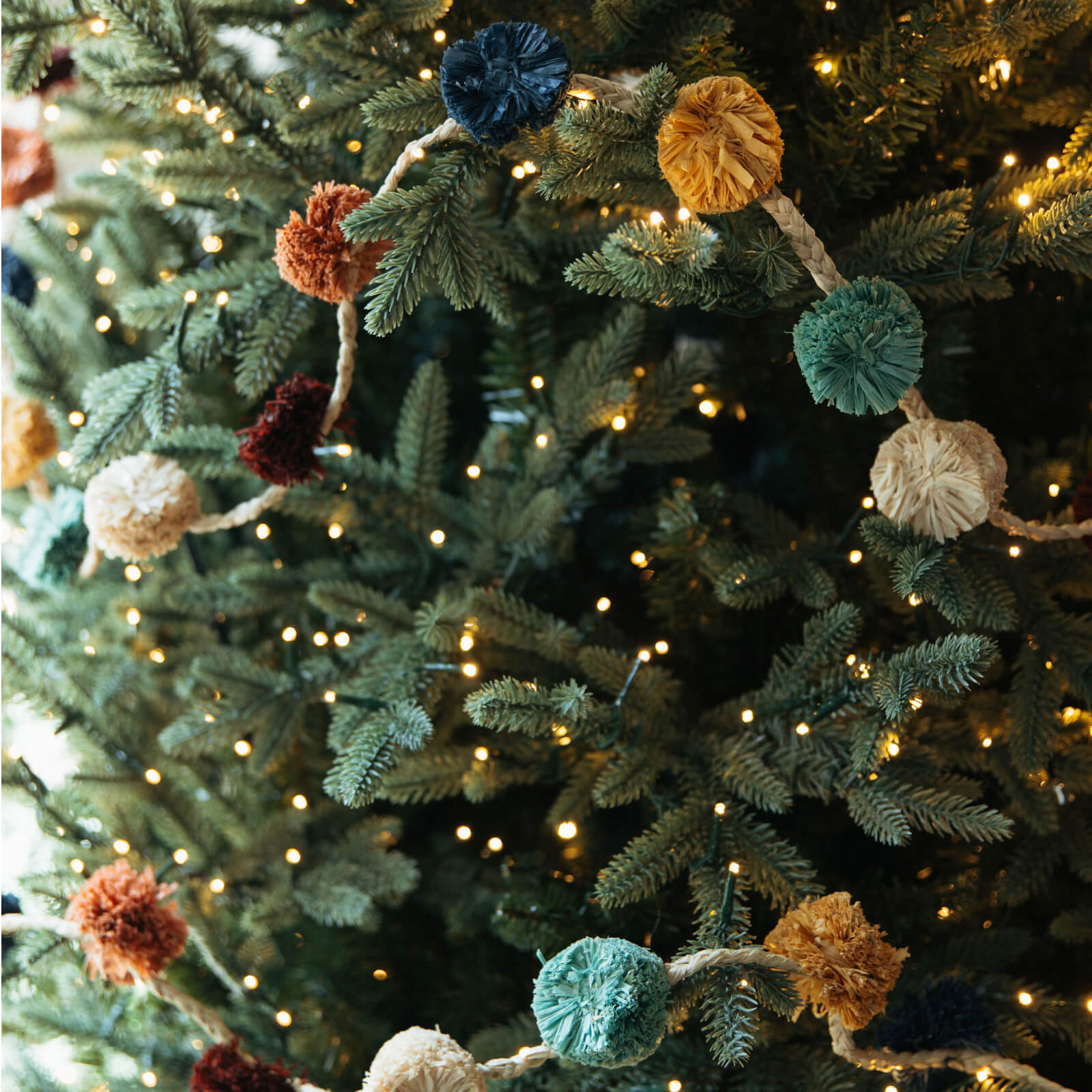 Decorative garland with colorful pom-poms on a Christmas tree with lights.