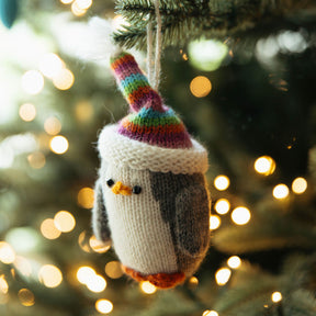 Knitted penguin ornament with a colorful hat hanging on a Christmas tree with blurred lights in the background.