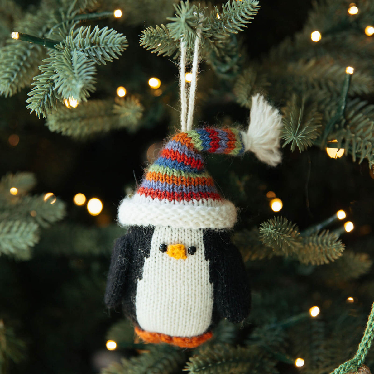 Knitted penguin ornament with a colorful hat hanging on a Christmas tree.