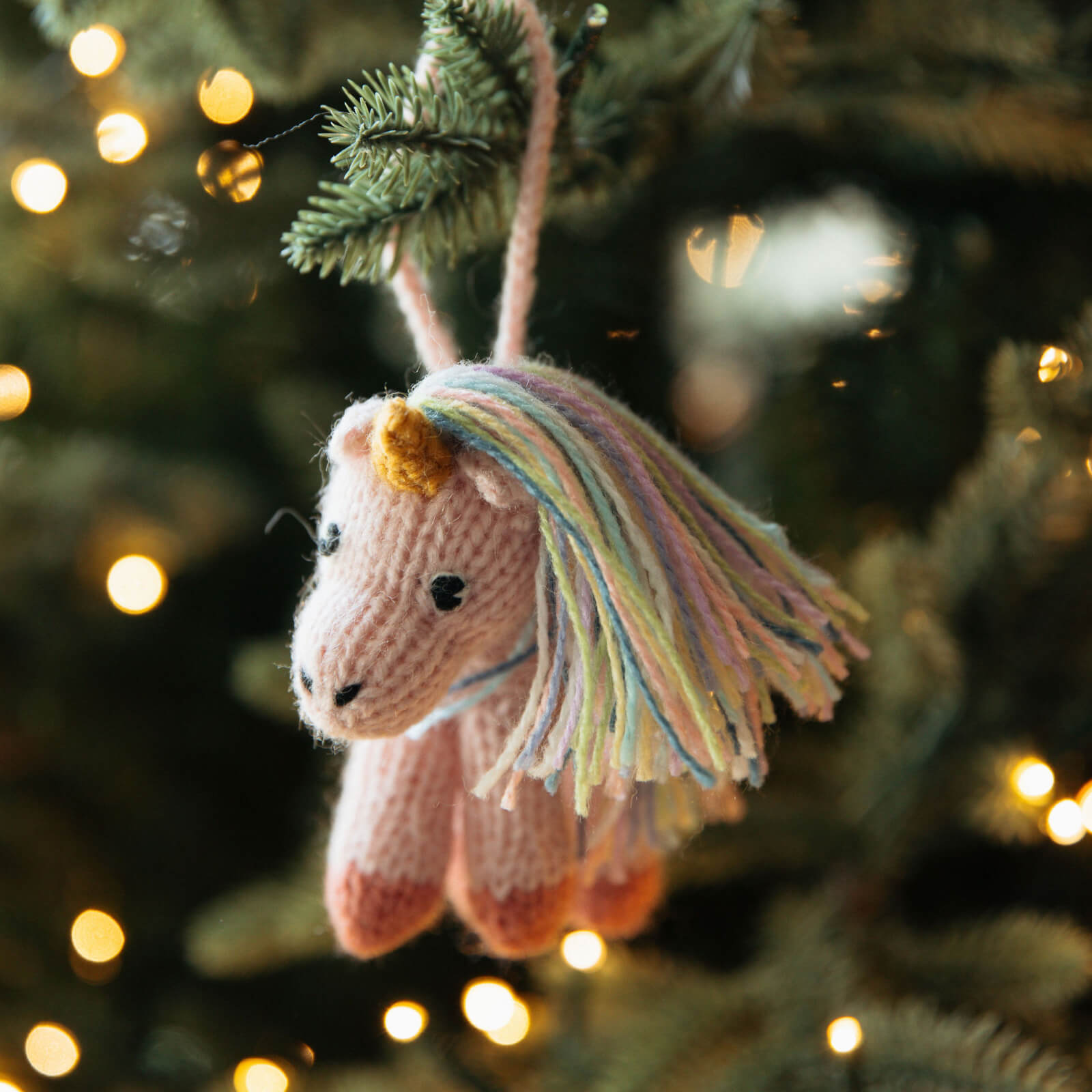Knitted unicorn ornament hanging on a Christmas tree with blurred lights in the background