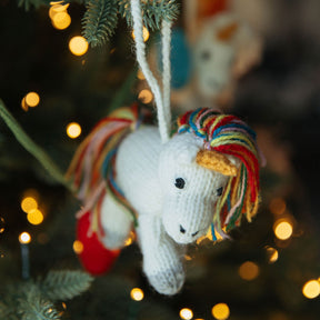 Knitted unicorn ornament with colorful mane hanging on a Christmas tree with blurred lights in the background.