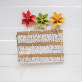 Three colorful paper flowers attached to a brown paper bag with a white pattern on a white wooden surface.
