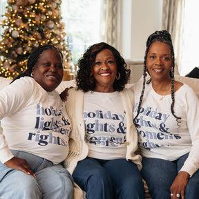 Three women sitting together wearing matching 'Holiday Lights & Memories' shirts in a festive setting.