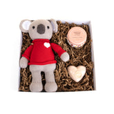 Knitted koala toy with a red sweater, candle, and heart-shaped object in a gift box.