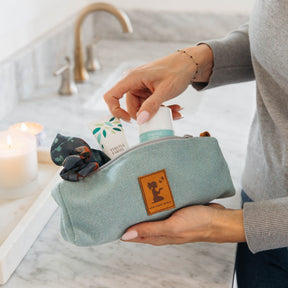 Person holding a light blue toiletry bag with products on a bathroom counter.