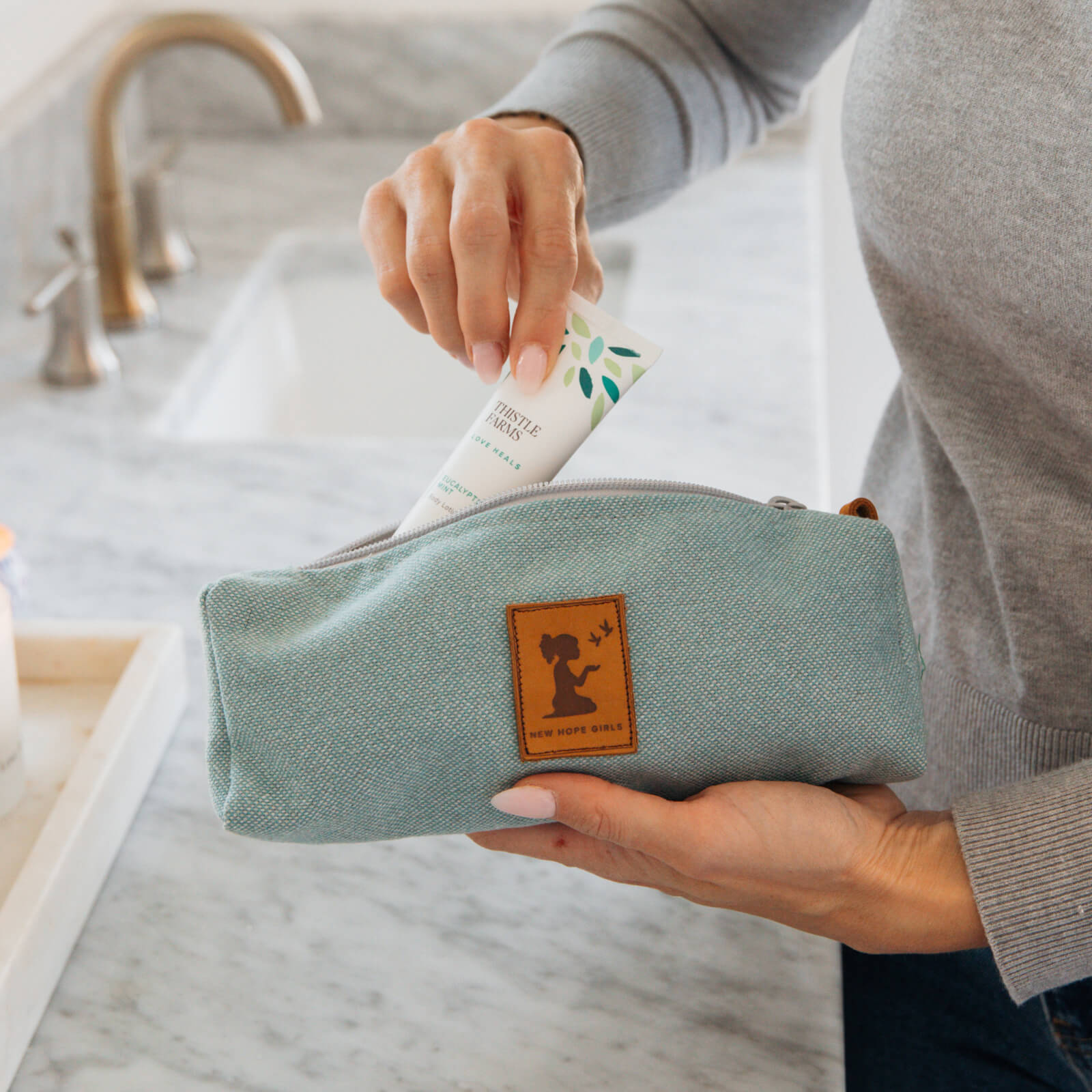 Person holding a light blue toiletry bag with a brand logo, with a bathroom setting in the background.