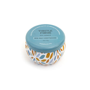 Candle jar with floral design and blue lid labeled 'Thistle Farms' on a white background