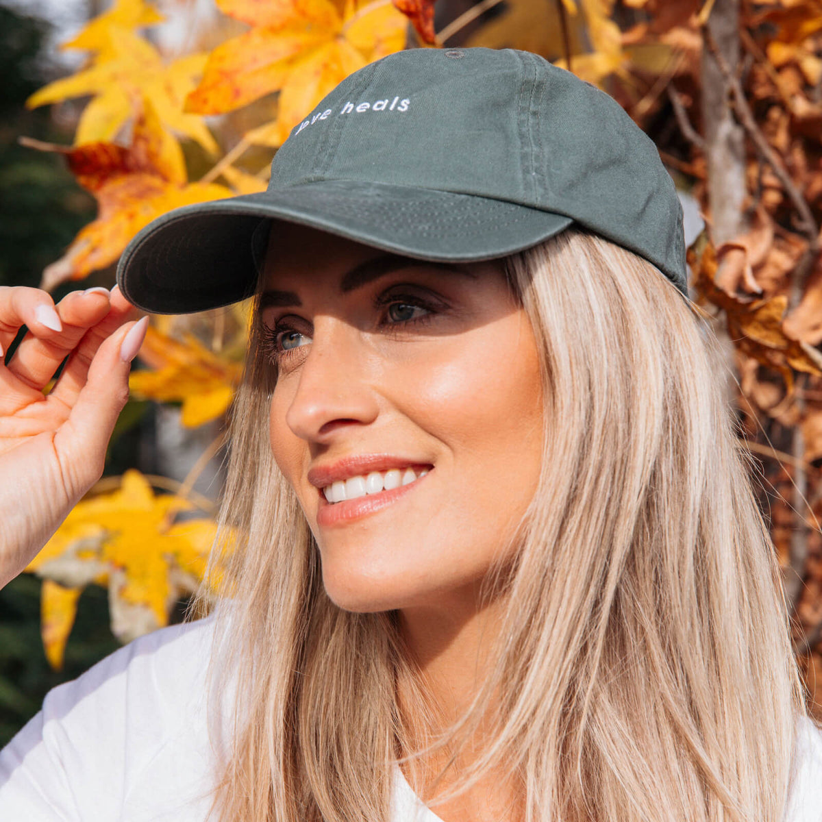 Woman wearing a green cap with text, standing in front of autumn leaves.