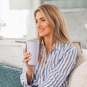 Woman holding a white tumbler with text, sitting on a couch in a cozy room.