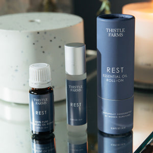 Thistle Farms Rest essential oil products on a reflective surface with a blurred background