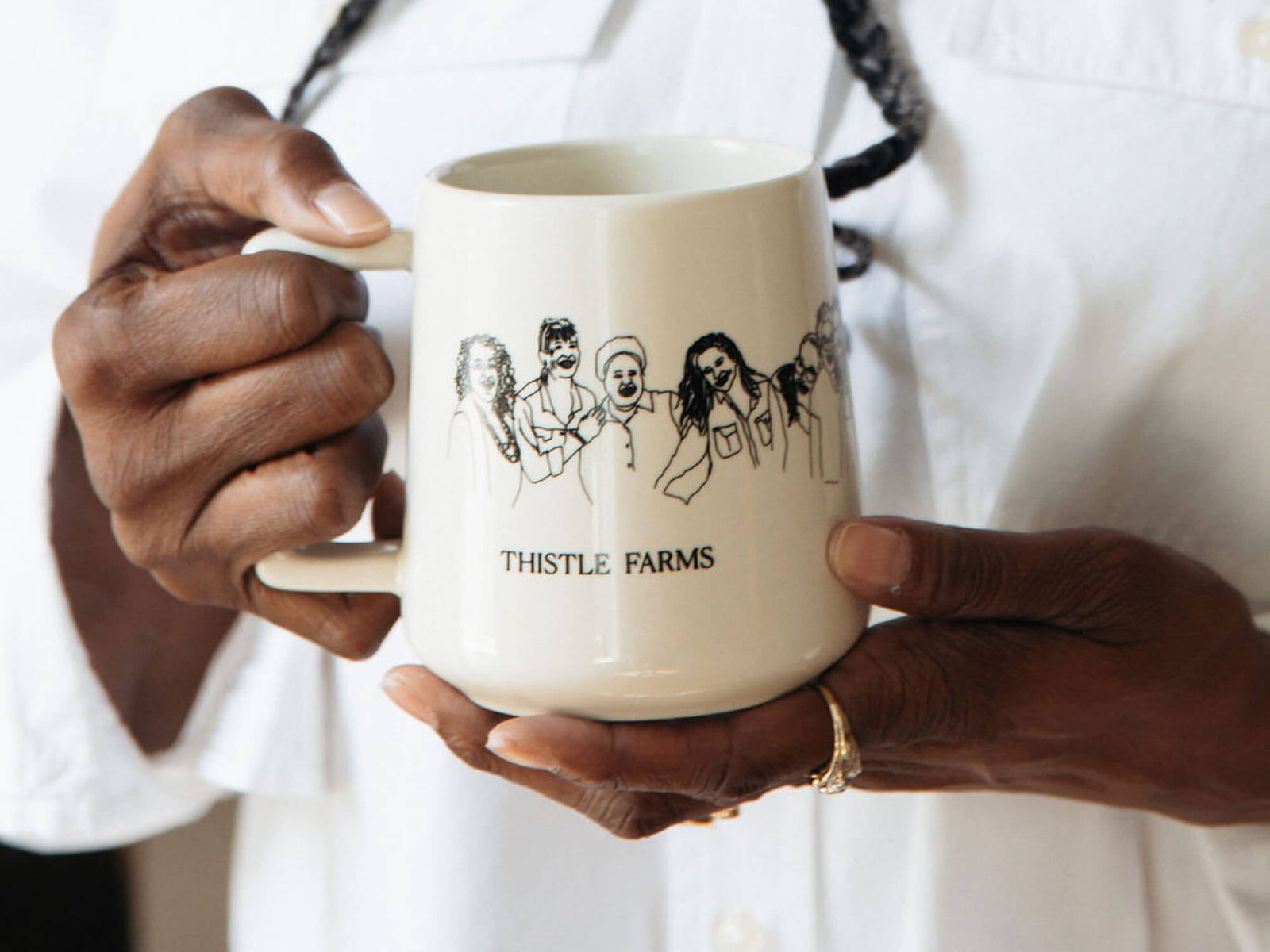 Person holding a mug with 'Thistle Farms' branding and sketch of the beautiful women of Thistle Farms.