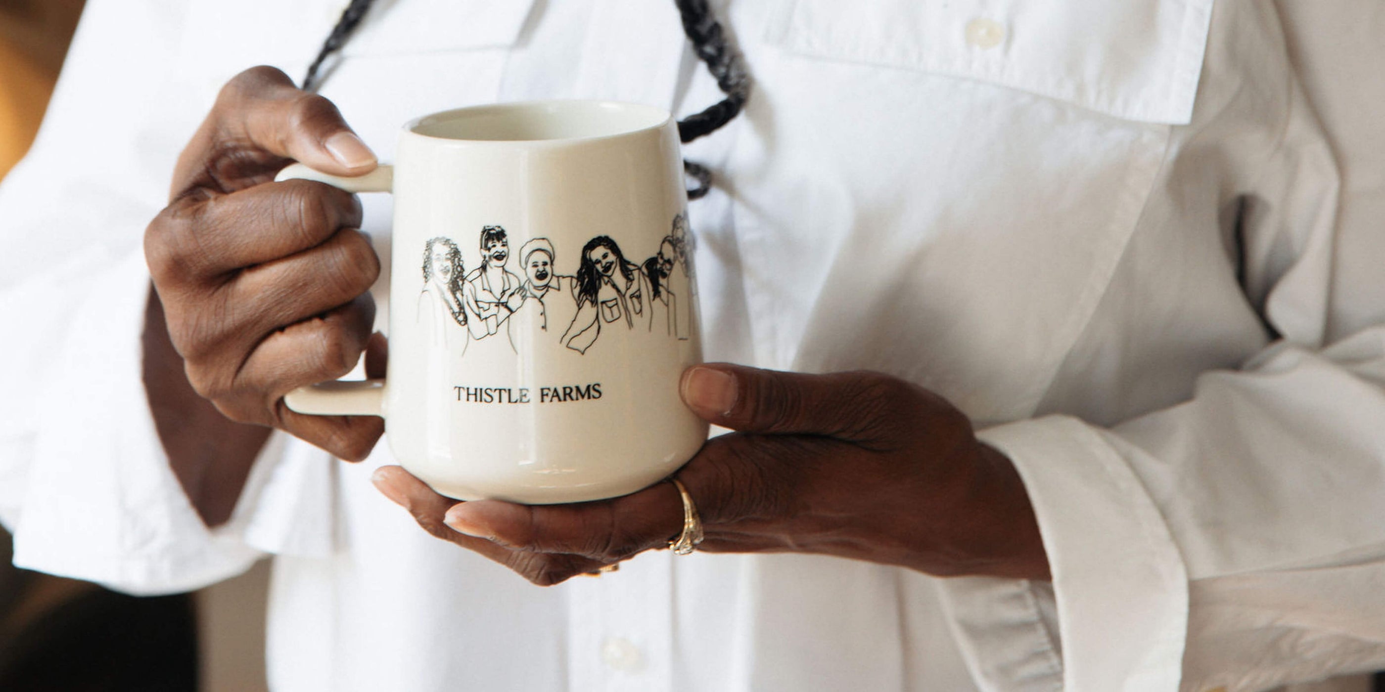 Person holding a mug with 'Thistle Farms' branding and a sketch of women hugging.