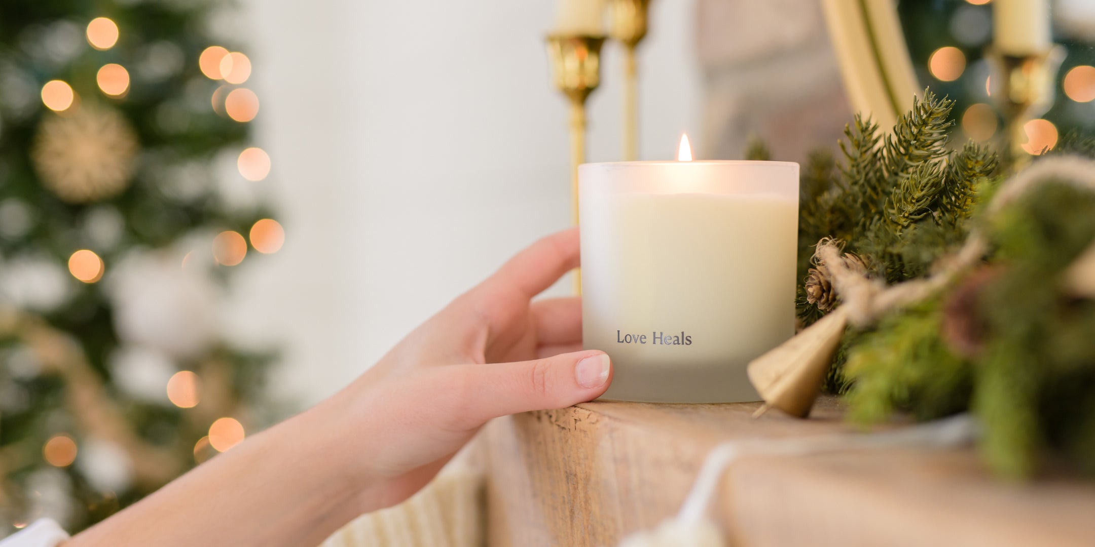 Hand holding a candle labeled 'Love Heals' with blurred Christmas decorations in the background