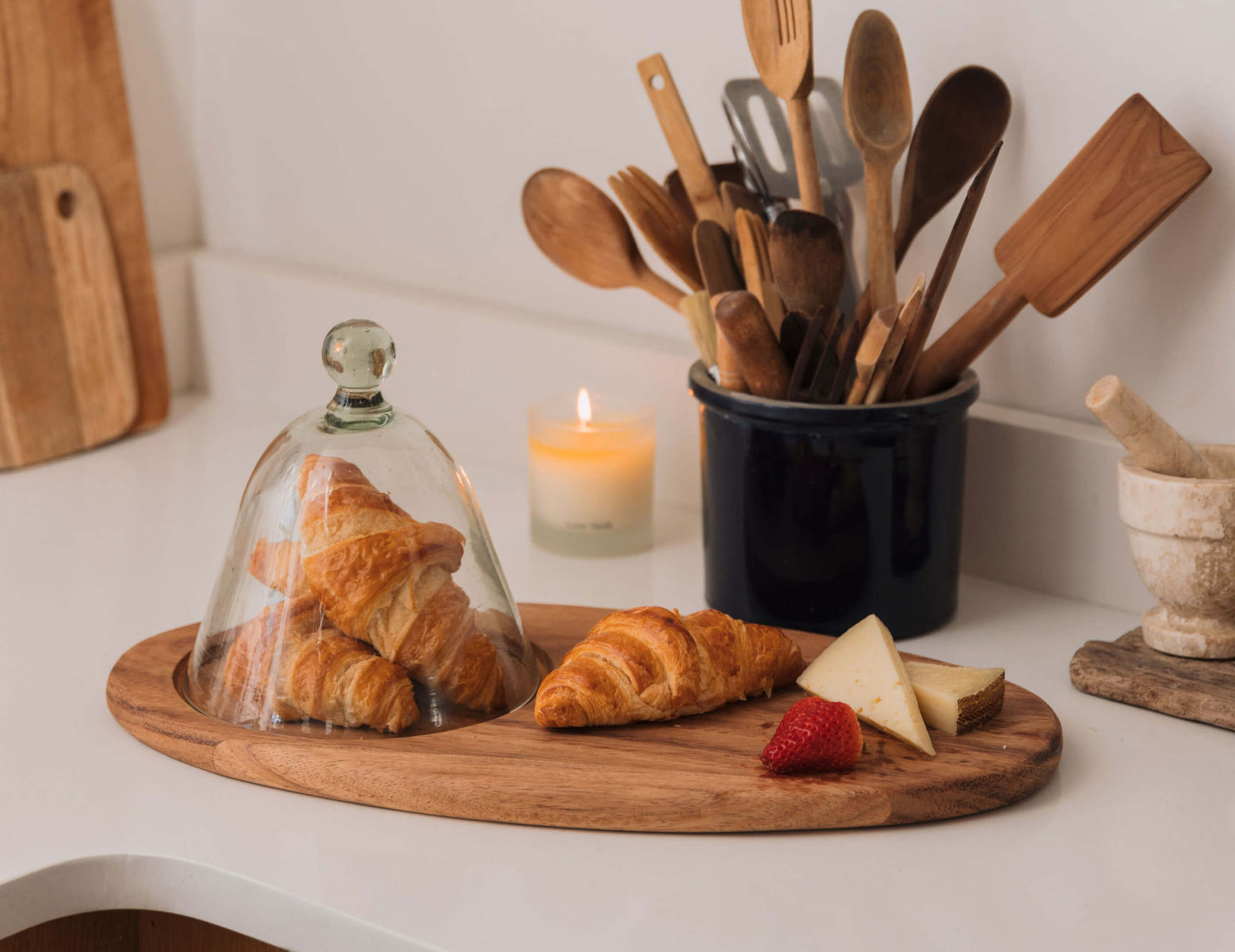 Wooden cutting board with croissants, a glass cloche, and kitchen utensils on a white surface.