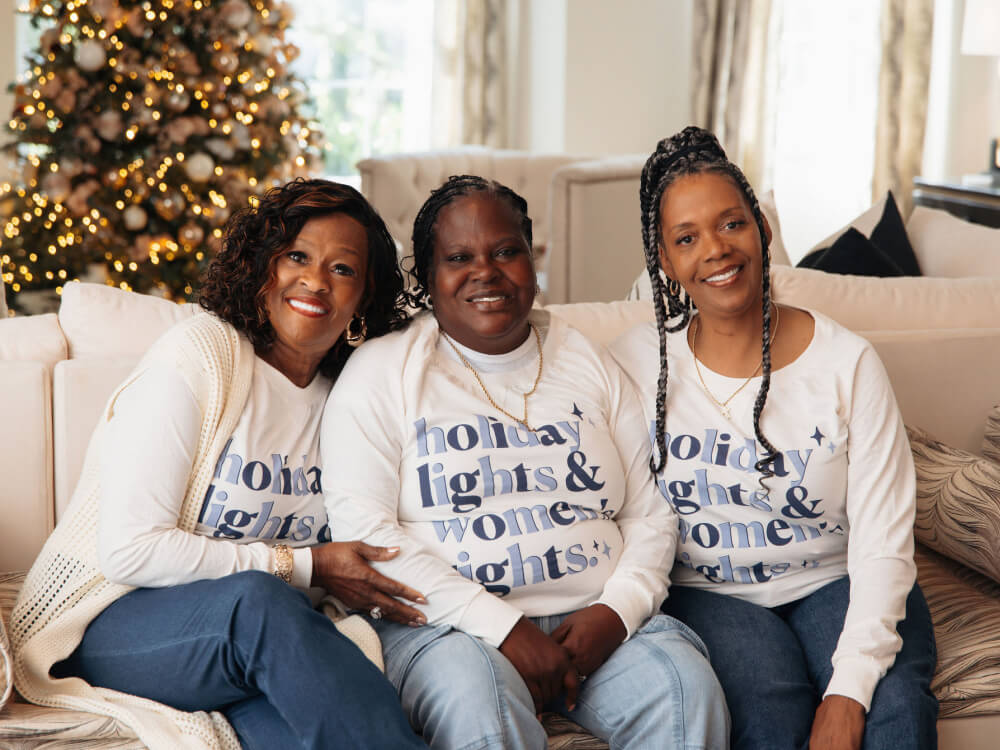 Three women sitting on a couch wearing matching holiday-themed shirts in a cozy living room.
