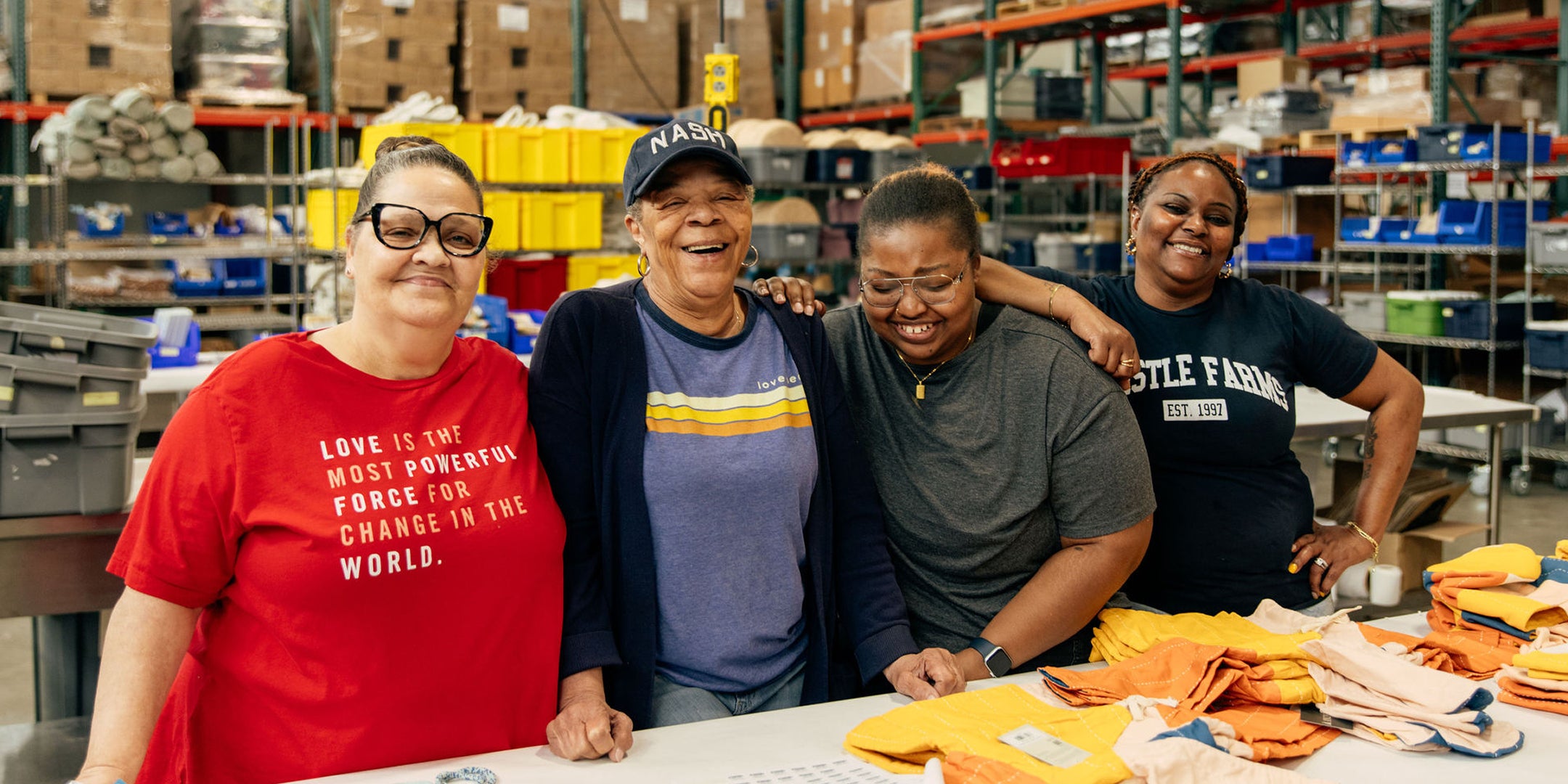 Four Thistle Farms employees standing together in a Thistle Farms Manufacturing Warehouse setting with tables and supplies in the background.