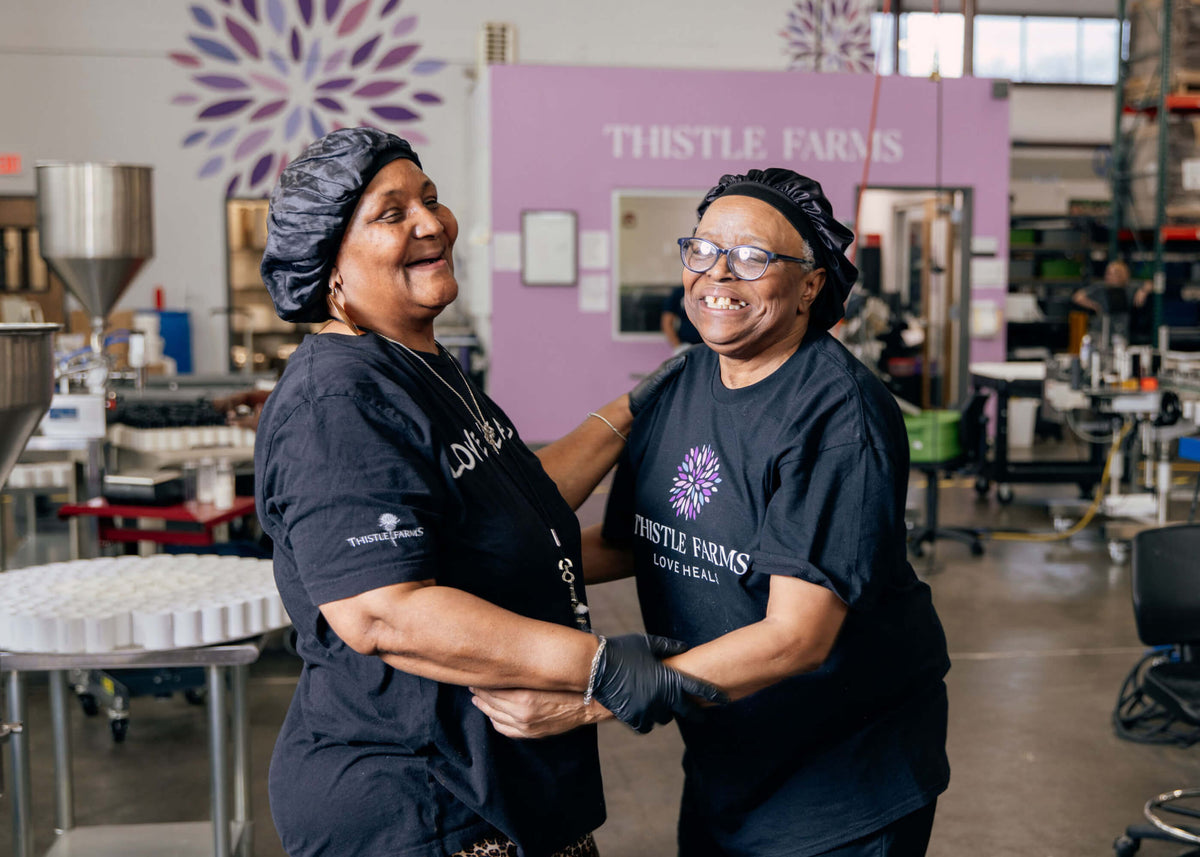 Two women in Thistle Farms shirts hugging in a warehouse setting