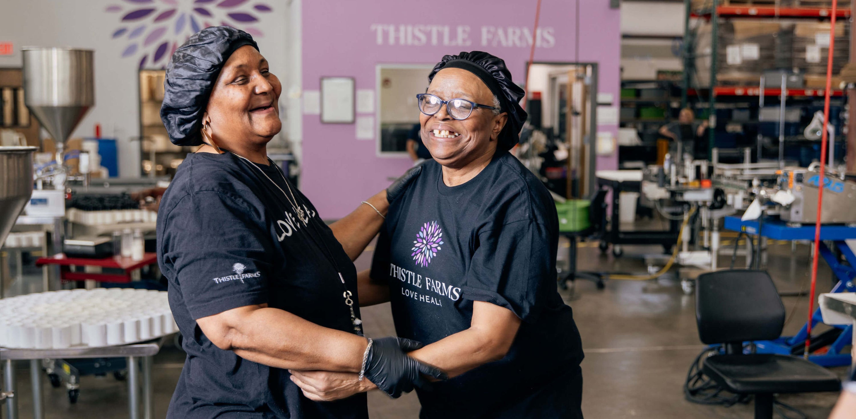 Two women in black shirts with 'Thistle Farms' logo in a warehouse setting.