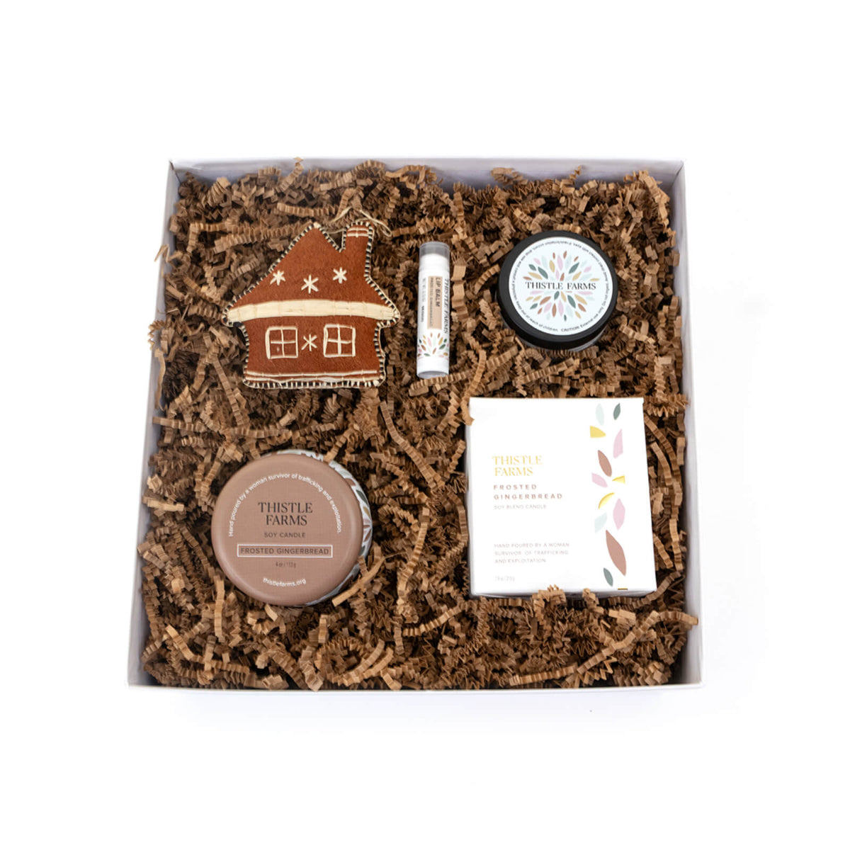 Gift set with Thistle Farms products including a house-shaped ornament, lip balm, and two containers on a white background.