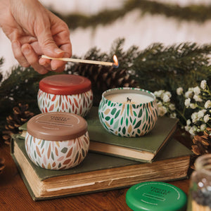 Hand lighting a candle with decorative lids on a stack of books, surrounded by greenery and flowers.