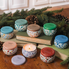 Assorted small candles with colorful lids on a wooden surface with books and greenery.