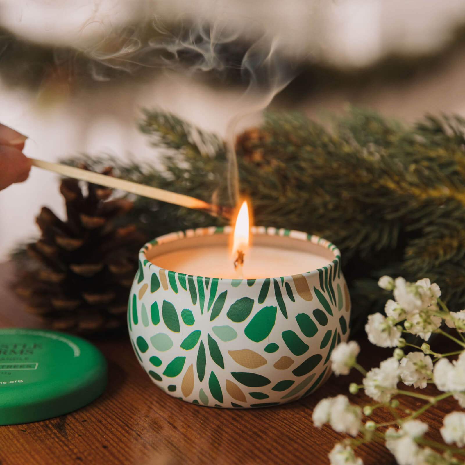 Candle with a pattern being lit next to pine cones and flowers on a wooden surface