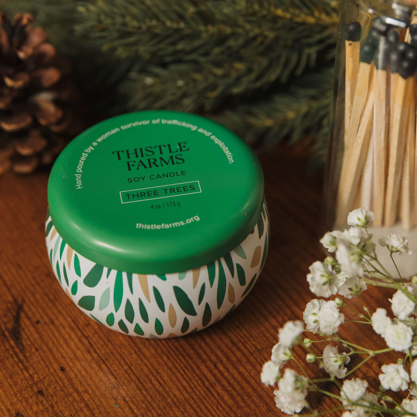 Thistle Farms soy candle jar with green lid on a wooden surface with decorative elements.