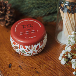 Thistle Farms candle with a red lid on a wooden surface with matches and flowers.