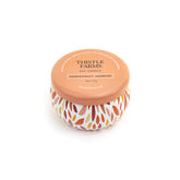 Thistle Farms soy candle with a leaf patterned base and orange lid on a white background