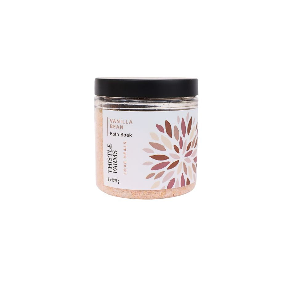 A jar of Parable Farms Love Reals Vanilla Bean Bath Soak on a solid white background. The product features a clear jar showing the light pink bath soak inside, a white label with a brown floral design, and a black lid.