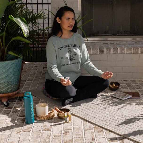 woman seated on yoga mat meditating near a Thistle Farms water bottle and candle